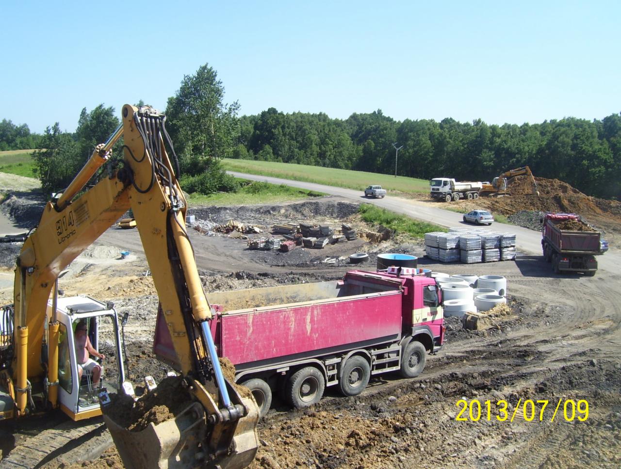 Żółta koparka Libronic 914 nabiera ziemię do czerwonej ciężarówki na placu budowy; w tle droga, drzewa, inne maszyny i elementy betonowe.