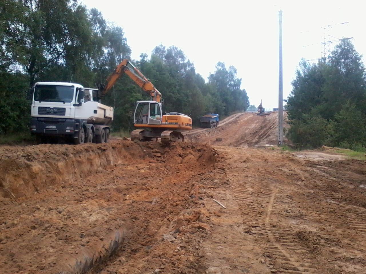 Żółta koparka Liebherr i biała ciężarówka MAN na placu budowy. W tle widać wykopany teren z drzewami i słupami energetycznymi. Prace ziemne w toku.
