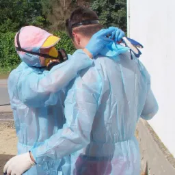 Dwie osoby w niebieskich kombinezonach ochronnych i maskach gazowych, obejmujące się na zewnątrz budynku. Jedna osoba ma różową czapkę z żółtym daszkiem, a druga ma niebieskie rękawice i trzyma...