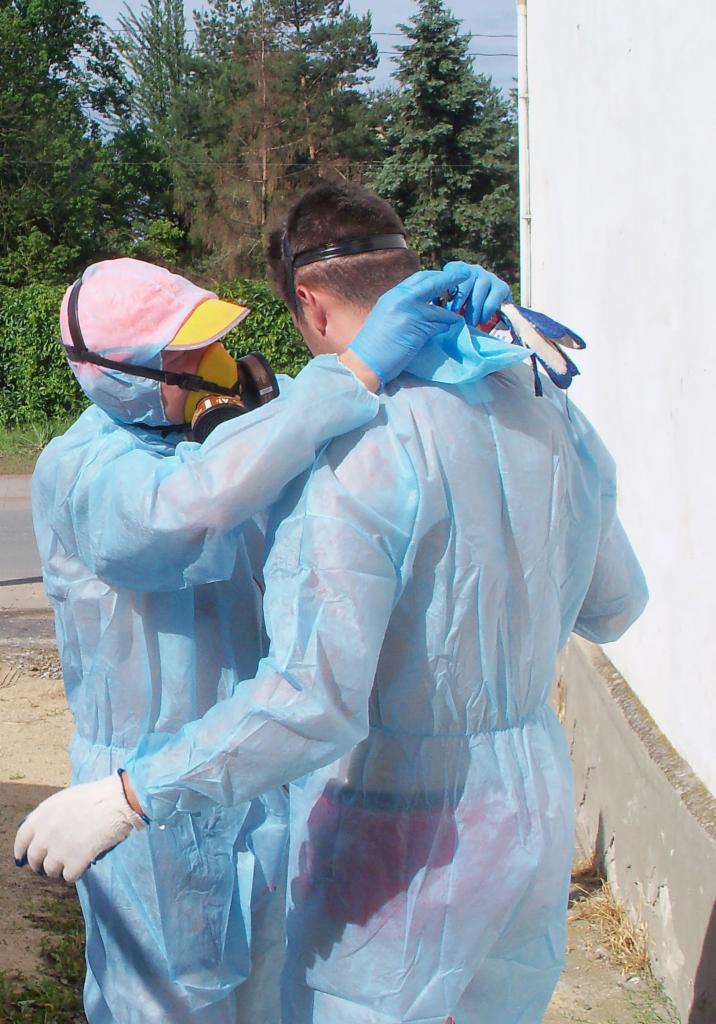 Dwie osoby w niebieskich kombinezonach ochronnych i maskach gazowych, obejmujące się na zewnątrz budynku. Jedna osoba ma różową czapkę z żółtym daszkiem, a druga ma niebieskie rękawice i trzyma...