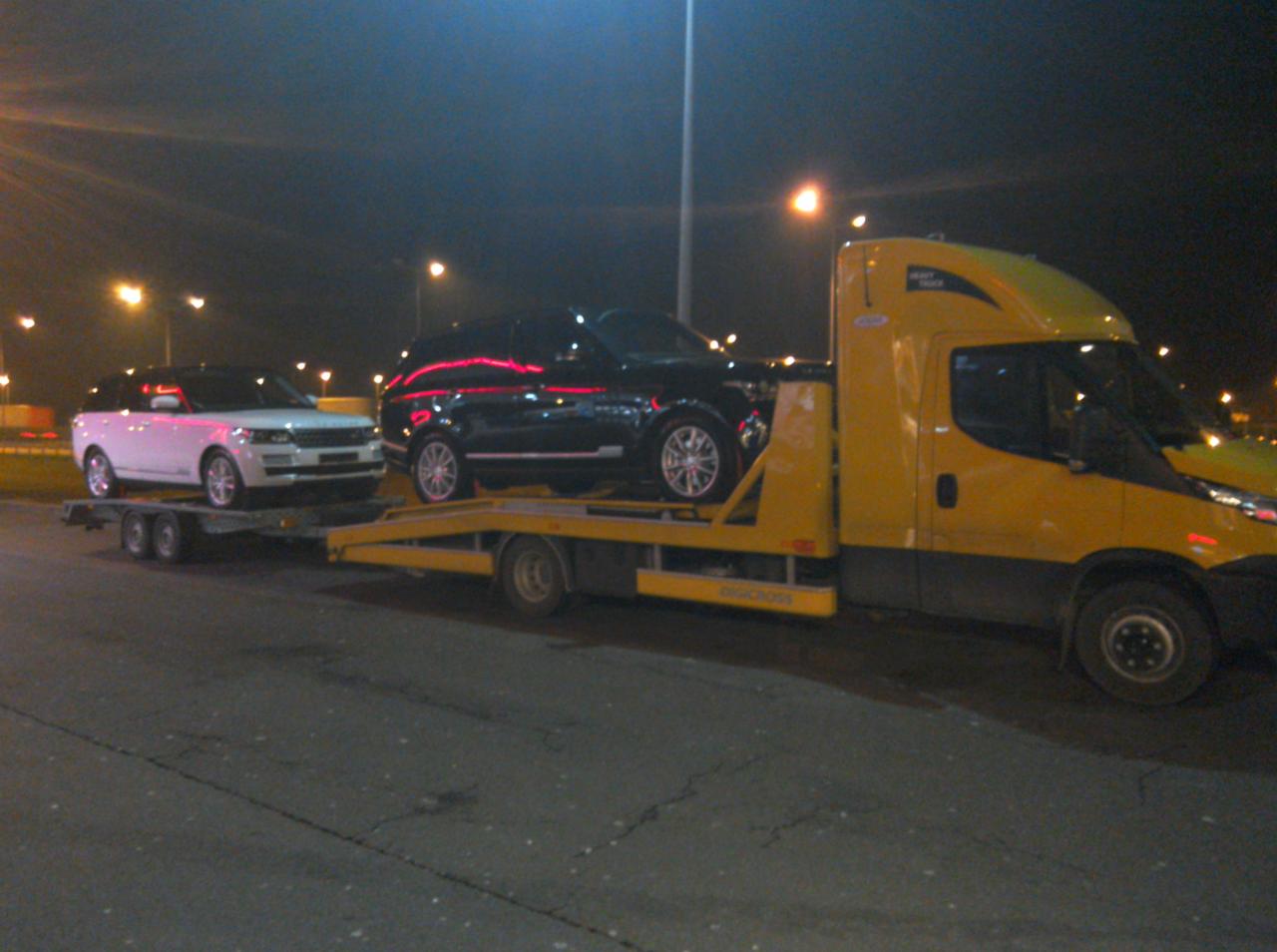 Żółta laweta z dwoma samochodami Range Rover (biały i czarny) na platformie, przewożona nocą na oświetlonym parkingu. Widoczne światła uliczne.