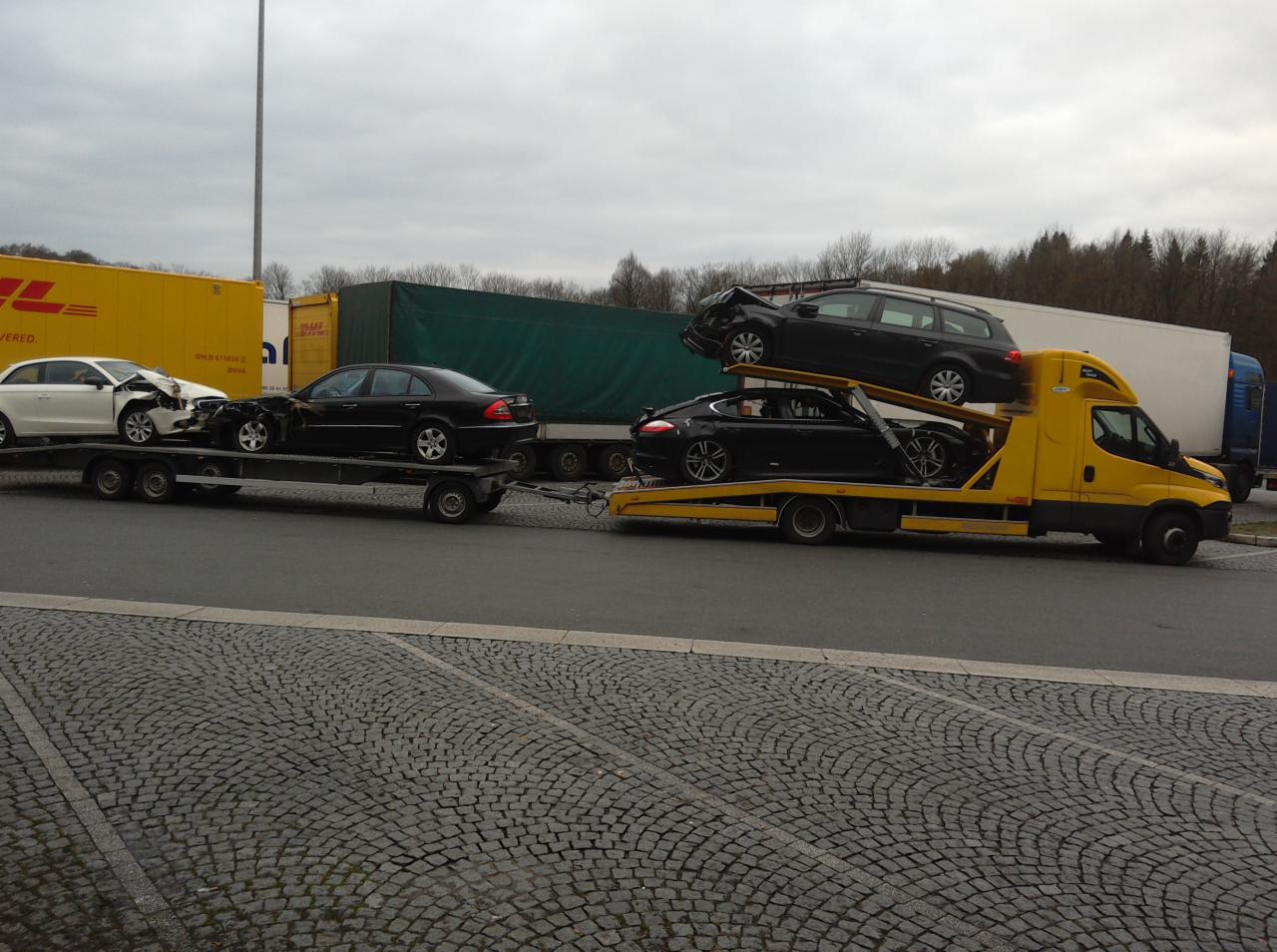 Żółta laweta przewozi dwa czarne samochody osobowe, a na przyczepie dwa kolejne, w tym jeden uszkodzony. W tle widoczne kontenery i drzewa.