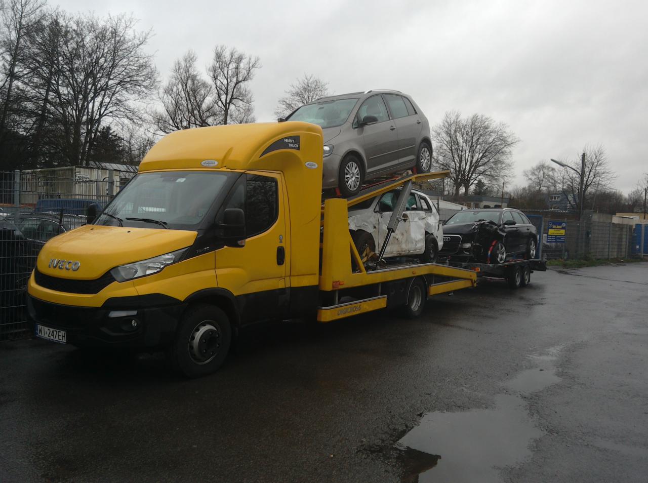 Żółta laweta Iveco z trzema uszkodzonymi autami na platformie, widok z boku, pochmurny dzień, asfaltowa nawierzchnia, drzewa w tle.