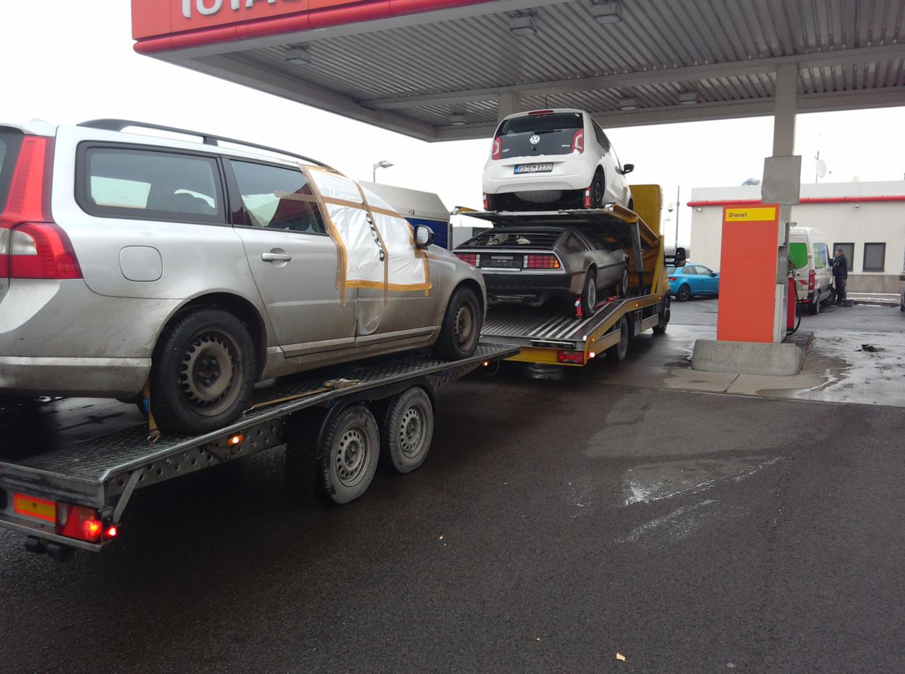 Laweta na stacji benzynowej z trzema samochodami: Volvo z uszkodzoną szybą, DeLorean i biały VW Up. Transport wielopoziomowy.