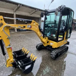 Żółta mini koparka CAT 301.6 z dwoma czarnymi łyżkami z żółtymi zębami stoi na mokrym, betonowym placu przed zadaszoną wiatą. Na łyżkach widoczne naklejki z kodami kreskowymi.