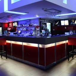 Nowoczesny bar z czerwonymi akcentami i podświetleniem LED. Krzesła barowe z czerwonymi siedziskami. W tle półki z alkoholem i dekoracyjne oświetlenie.