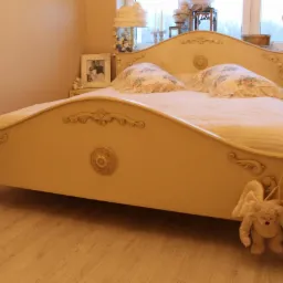 Eleganckie łóżko w stylu retro z dekoracyjnymi zdobieniami i okrągłymi motywami na tle stonowanej sypialni. Uwagę przykuwa pluszowy aniołek zawieszony na boku łóżka.