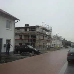 Rozbudowa domu w trakcie prac: budynek w rusztowaniach, częściowo otynkowany, widoczny z ulicy z zaparkowanymi autami.