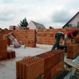 Wznoszenie murów parteru domu z czerwonej cegły ceramicznej Porotherm; dwóch murarzy przy pracy, jeden nakłada zaprawę, drugi układa cegły; widoczne sznurki wyznaczające linię muru, palety...