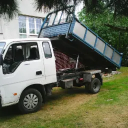 Biała wywrotka Kia K2700 z podniesioną, błękitną skrzynią ładunkową na tle zielonego trawnika i budynku. Widoczne detale konstrukcyjne pojazdu.