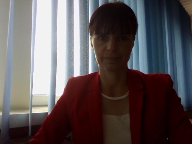 Portret kobiety w czerwonej marynarce na tle okna z żaluzjami w Lublinie. Ujęcie od przodu, widoczna górna część ciała. Neutralny wyraz twarzy.