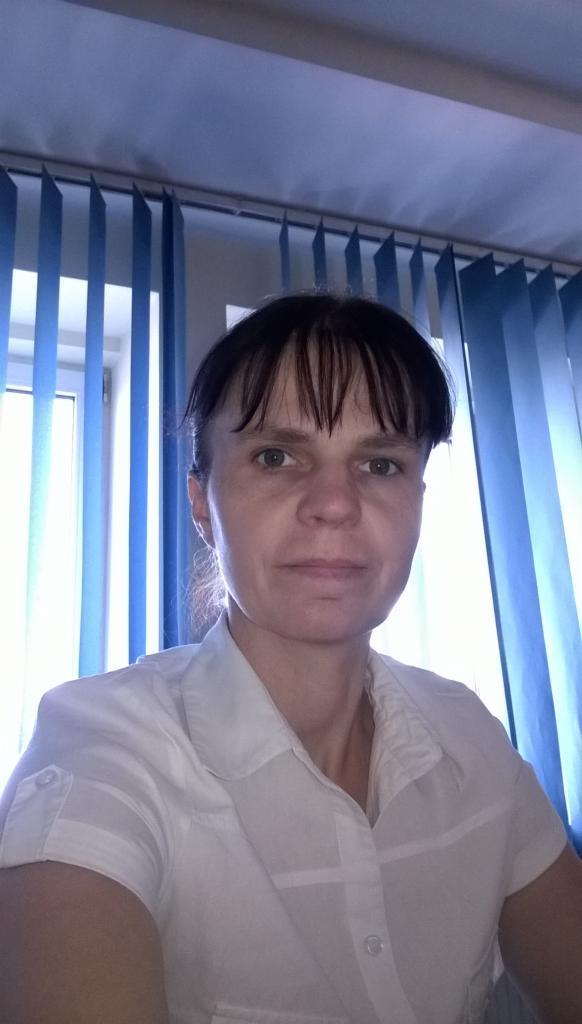 Portret kobiety w białej koszuli na tle pionowych, niebieskich żaluzji. Kadr od ramion w górę, oświetlenie dzienne, neutralny wyraz twarzy.