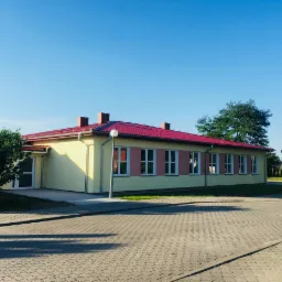 Nowo wybudowany budynek z żółtą elewacją i czerwonym dachem, widoczny z zewnątrz na tle błękitnego nieba i zieleni drzew.