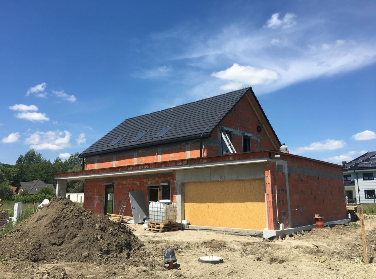 Dom w stanie surowym otwartym z ciemną dachówką, widoczne okna dachowe, fragment nieotynkowanej ściany z cegły oraz sterta ziemi na pierwszym planie.