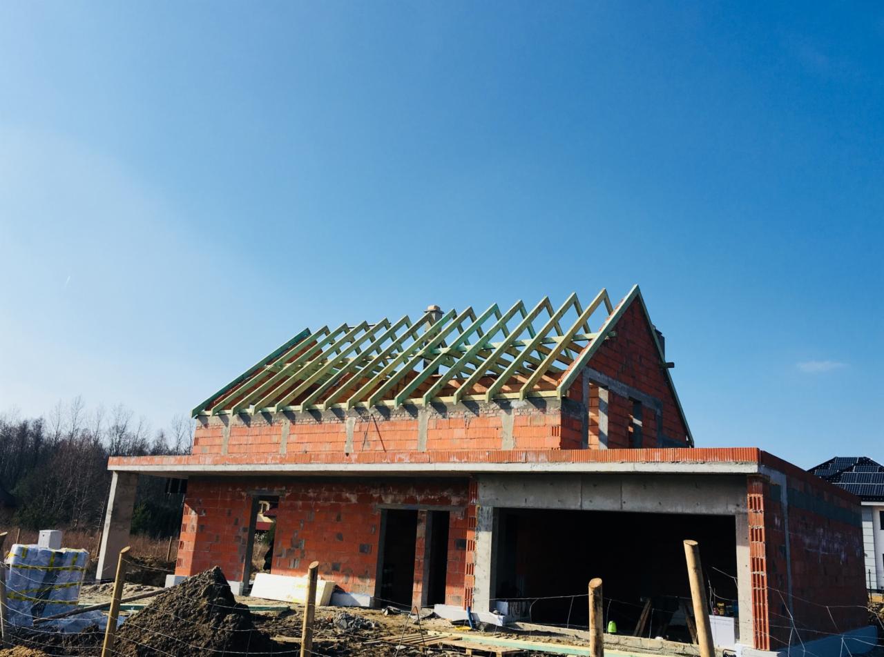 Surowy stan budynku jednorodzinnego z częściowo wykonaną konstrukcją drewnianą dachu, widoczne cegły i bloczki, w tle błękitne niebo.