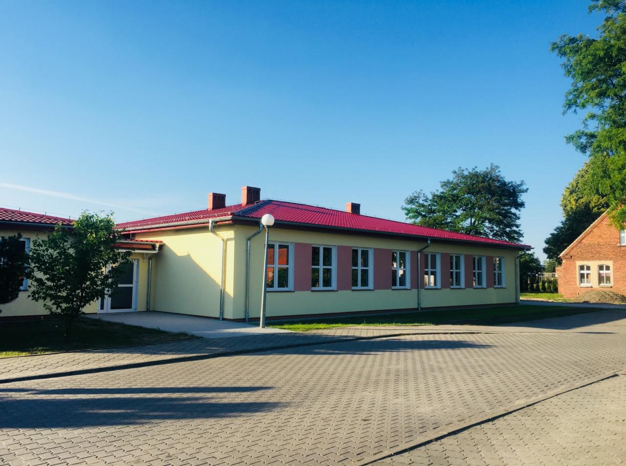 Nowo wybudowany budynek z żółtą elewacją i czerwonym dachem, widoczny z zewnątrz na tle błękitnego nieba i zieleni drzew.