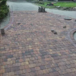 Ułożona kostka brukowa w odcieniach brązu i beżu, tworząca ścieżkę z delikatnym łukiem. W tle widoczny budynek gospodarczy i trawnik. Częściowo ułożona obwódka z kostki.