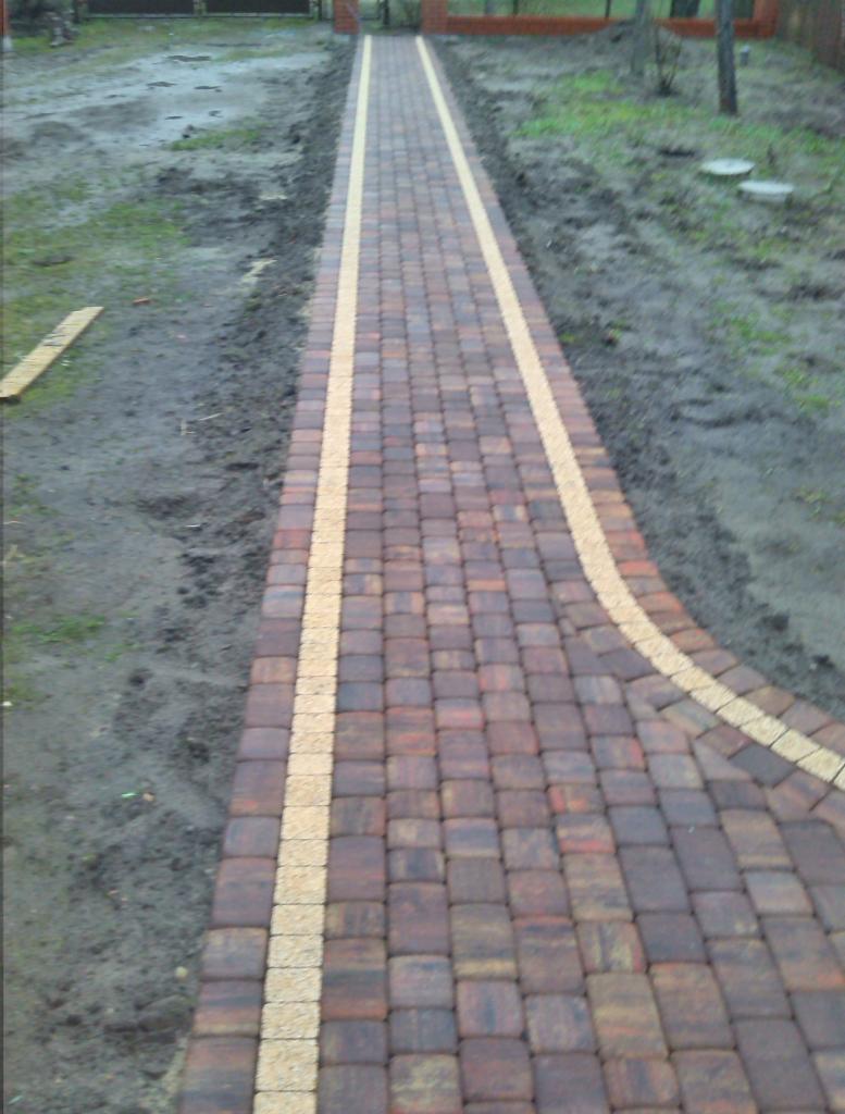 Ścieżka z kostki brukowej w odcieniach brązu, wykończona jasnymi pasami po bokach, prowadząca przez teren z ziemią i trawą w kierunku muru z cegły.