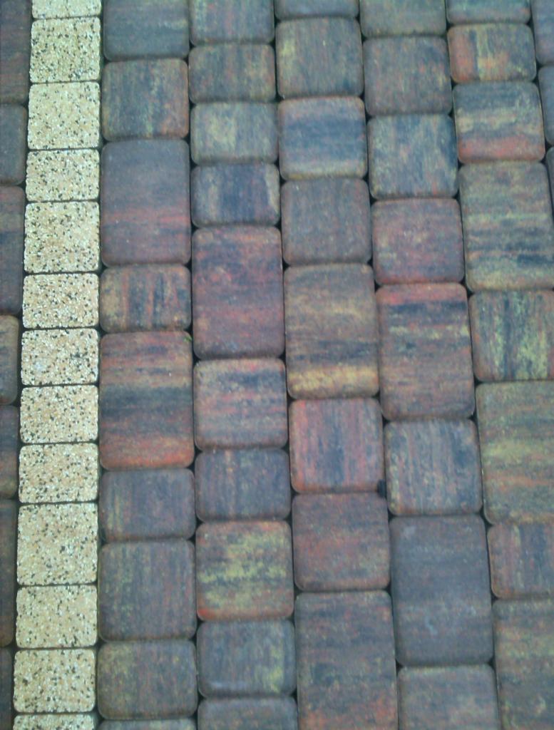 Ułożona kostka brukowa w odcieniach brązu i beżu, tworząca regularny wzór. Jasna linia kostki brzegowej kontrastuje z ciemniejszą, wzorzystą powierzchnią.