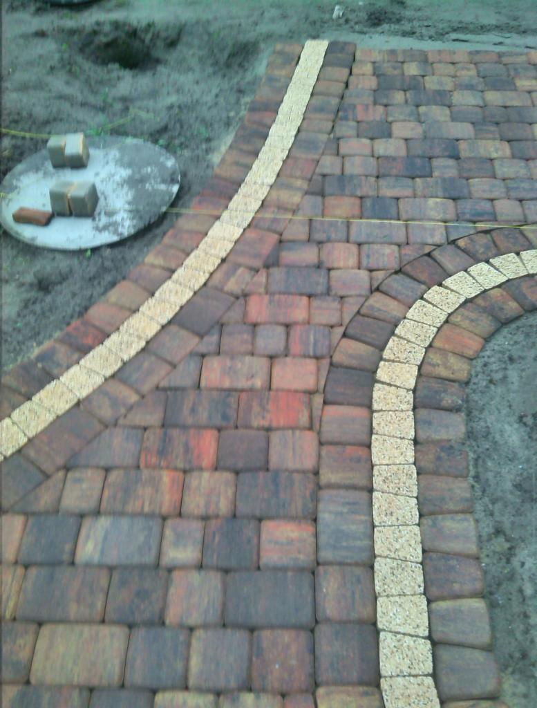 Ułożona, wzorzysta kostka brukowa w odcieniach brązu i beżu, z elementami dekoracyjnymi. Widoczne narzędzia brukarskie i fragment terenu przed zagospodarowaniem.