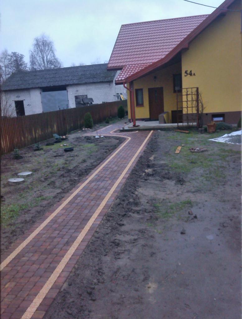 Kręta ścieżka z czerwonej kostki brukowej z jasnymi obrzeżami prowadzi do żółtego domu z czerwonym dachem. Widoczne świeżo posadzone rośliny i błotniste podłoże.