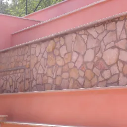 Różowa ściana z kamienną mozaiką, wykończenia z jasnego kamienia. Nietypowe połączenie kolorów i faktur, budynek w słoneczny dzień.