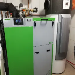 Nowoczesny, dwuczłonowy piec na paliwo stałe z zielonym panelem bocznym i czarnym sterownikiem elektronicznym, obok zasobnik ciepłej wody użytkowej Brötje z czarną obudową izolacyjną, na podłodze...