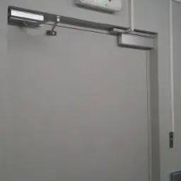 Szare drzwi z samozamykaczem i lampą awaryjną z zielonym światłem. Widoczne elementy montażowe i fragment ściany. Kadr statyczny, oświetlenie równomierne.