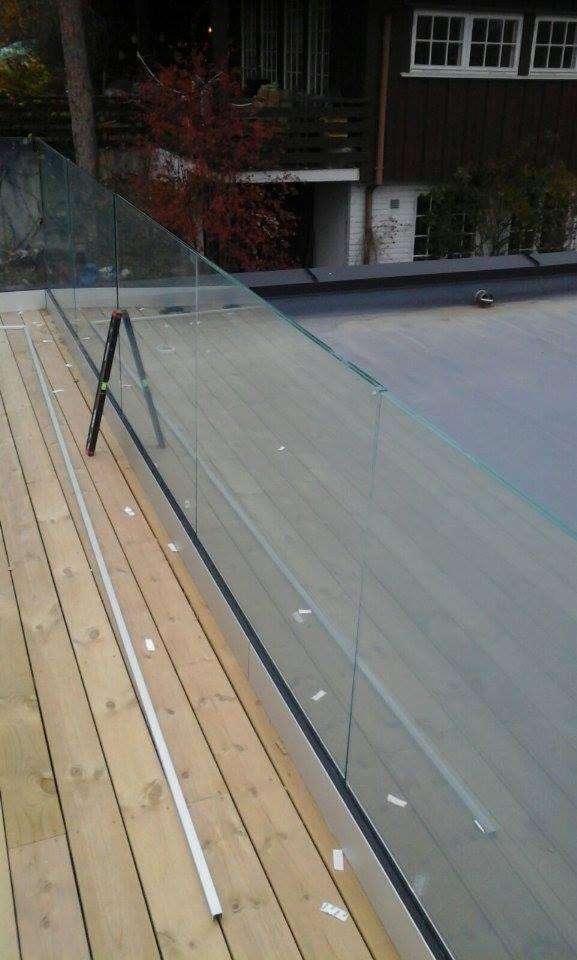 Szklana balustrada tarasu z drewnianą podłogą, poziomica oparta o szkło, widok na budynek mieszkalny i drzewo z czerwonymi liśćmi w tle.
