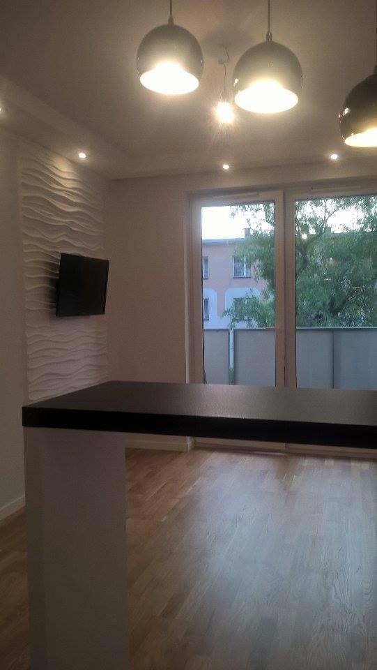 Nowoczesne wnętrze: dekoracyjna ściana z falistą strukturą, wiszący telewizor, drewniana podłoga w jodełkę, czarny blat i designerskie lampy sufitowe.