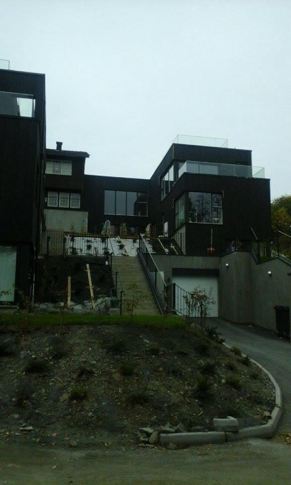 Nowoczesny dom w trakcie ocieplania, widoczne rusztowania i zabezpieczenia na tarasie oraz elewacji, teren wokół domu z nasadzeniami, pochmurne niebo.