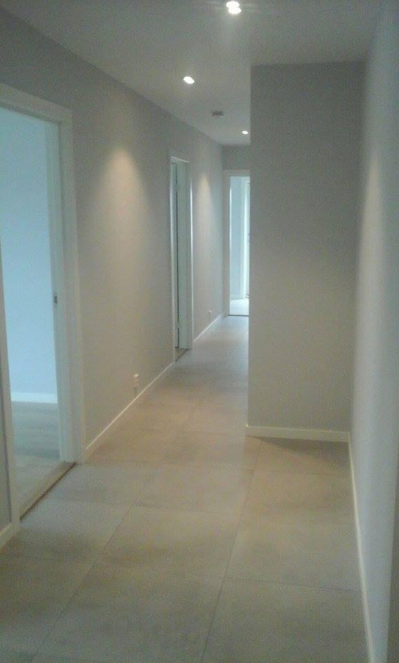 Długi korytarz w odcieniach szarości z białymi drzwiami i podłogą z płytek, oświetlony punktowo w suficie.