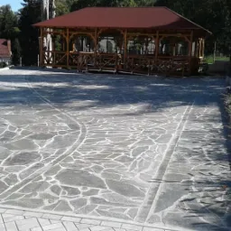 Szara, nieregularna kamienna nawierzchnia z widocznymi śladami po okręgu, obramowana kostką granitową, prowadząca do drewnianej altany na tle zieleni.