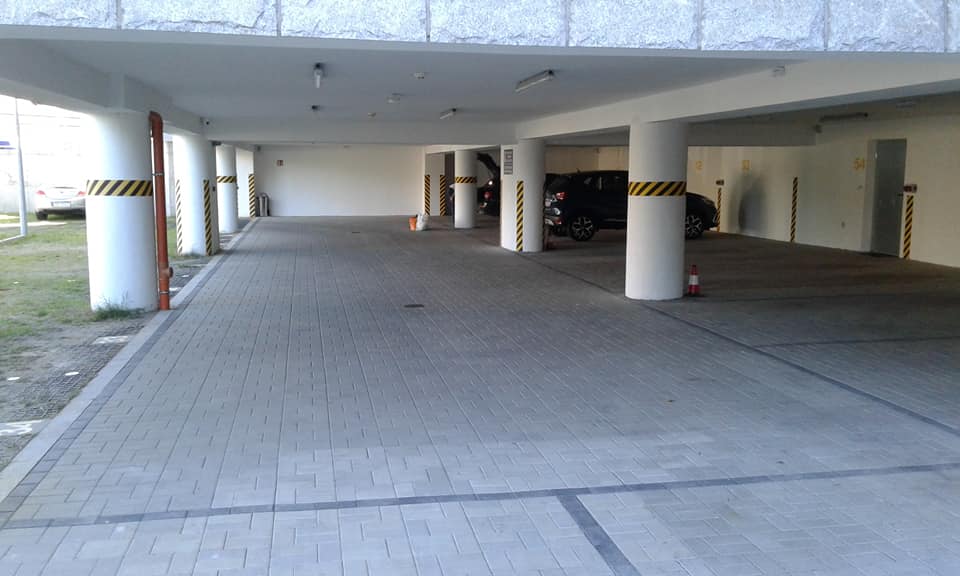 Zadaszony parking z szarej kostki brukowej ułożonej we wzór jodełki, białe filary z żółto-czarnymi pasami ostrzegawczymi, zaparkowane samochody w tle.