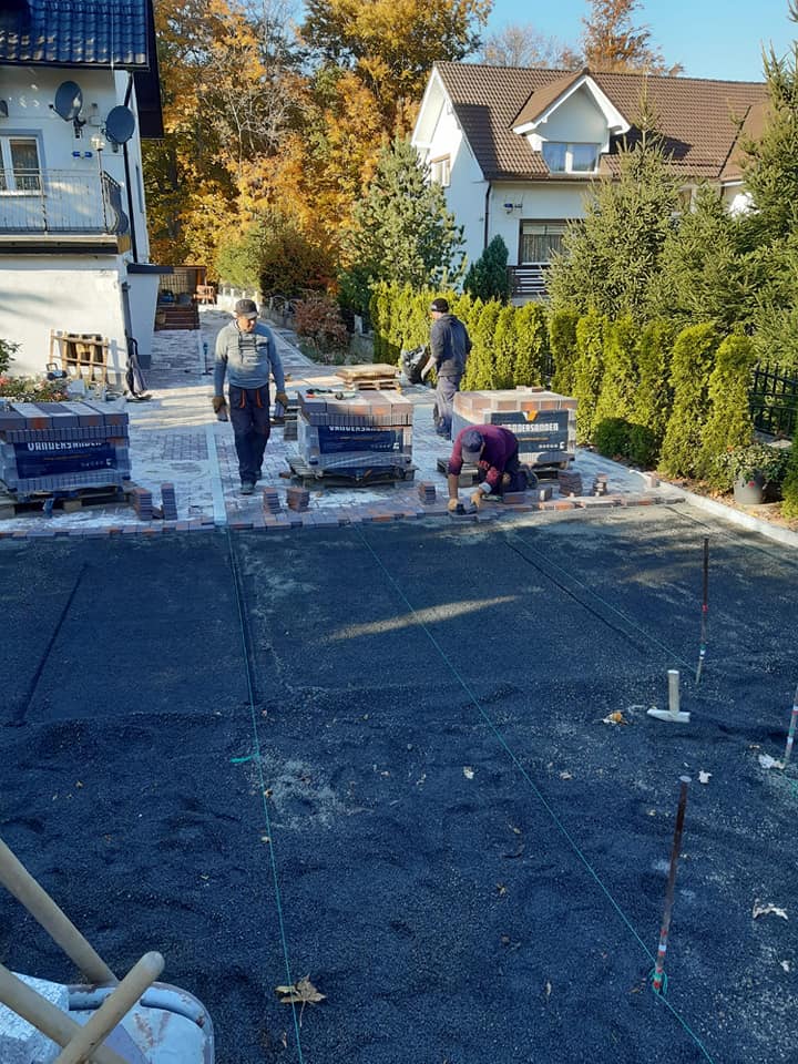 Prace brukarskie w Świeradowie-Zdroju: ekipa układa czerwoną kostkę brukową na przygotowanym podłożu, widoczne palety z kostką i narzędzia.