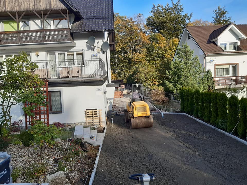 Żółty walec drogowy ubija podłoże pod przyszłą nawierzchnię z kostki, widoczne palety z kostką, dom z balkonem i jesienne drzewa w tle.