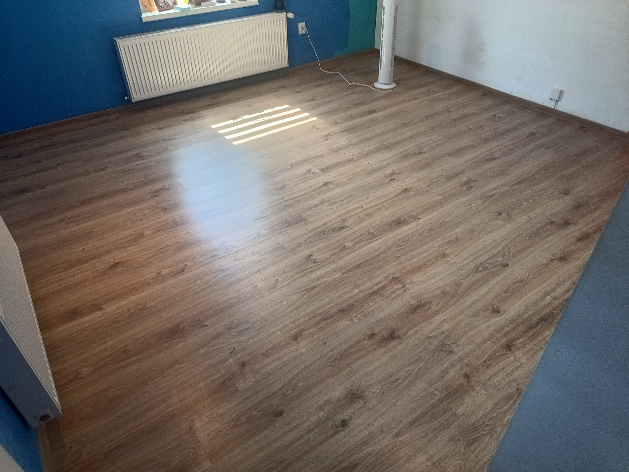 Ułożone panele podłogowe w odcieniach drewna, widoczne promienie słoneczne na powierzchni, niebieska ściana w tle.