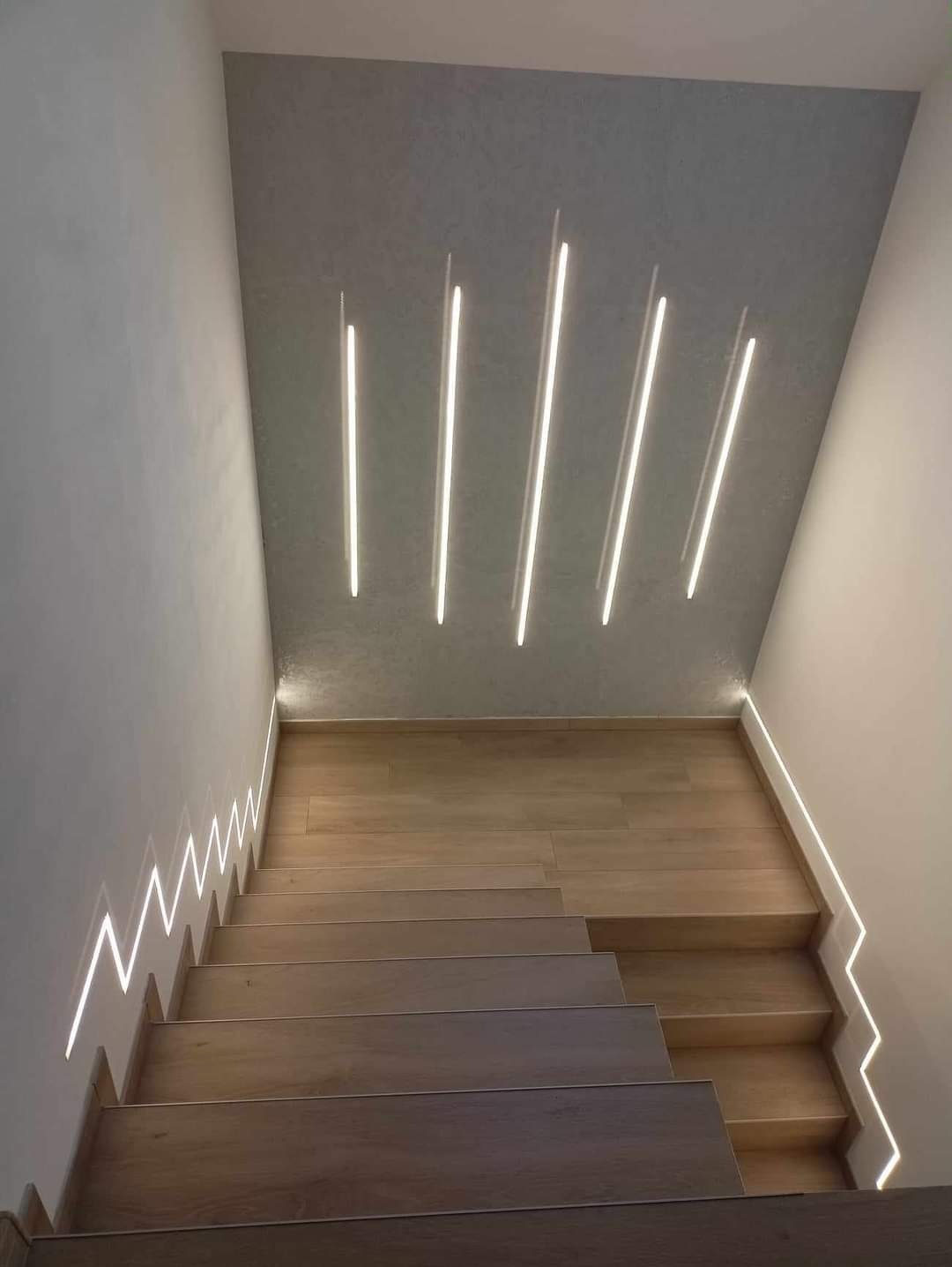 Nowoczesne schody z drewnianymi stopniami i oświetleniem LED wzdłuż ściany i na suficie, tworzące unikalny wzór świetlny.