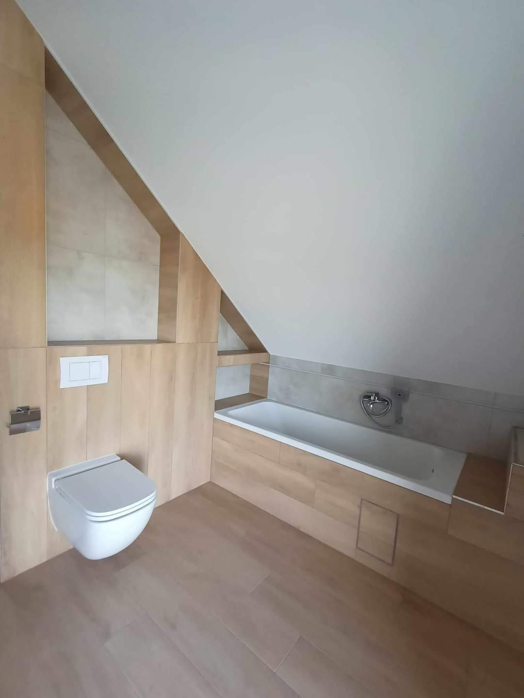 Wykończona łazienka na poddaszu ze skośnym sufitem, z drewnopodobnymi płytkami na ścianach i podłodze, wisząca toaleta z białym przyciskiem spłukującym, biała wanna obudowana płytkami i chromowana...