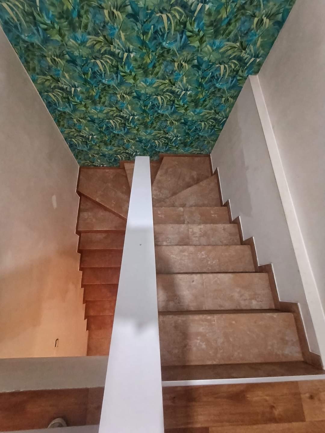 Schody wyłożone brązowymi płytkami, biała balustrada, sufit z tapetą w tropikalne liście, widok z góry.