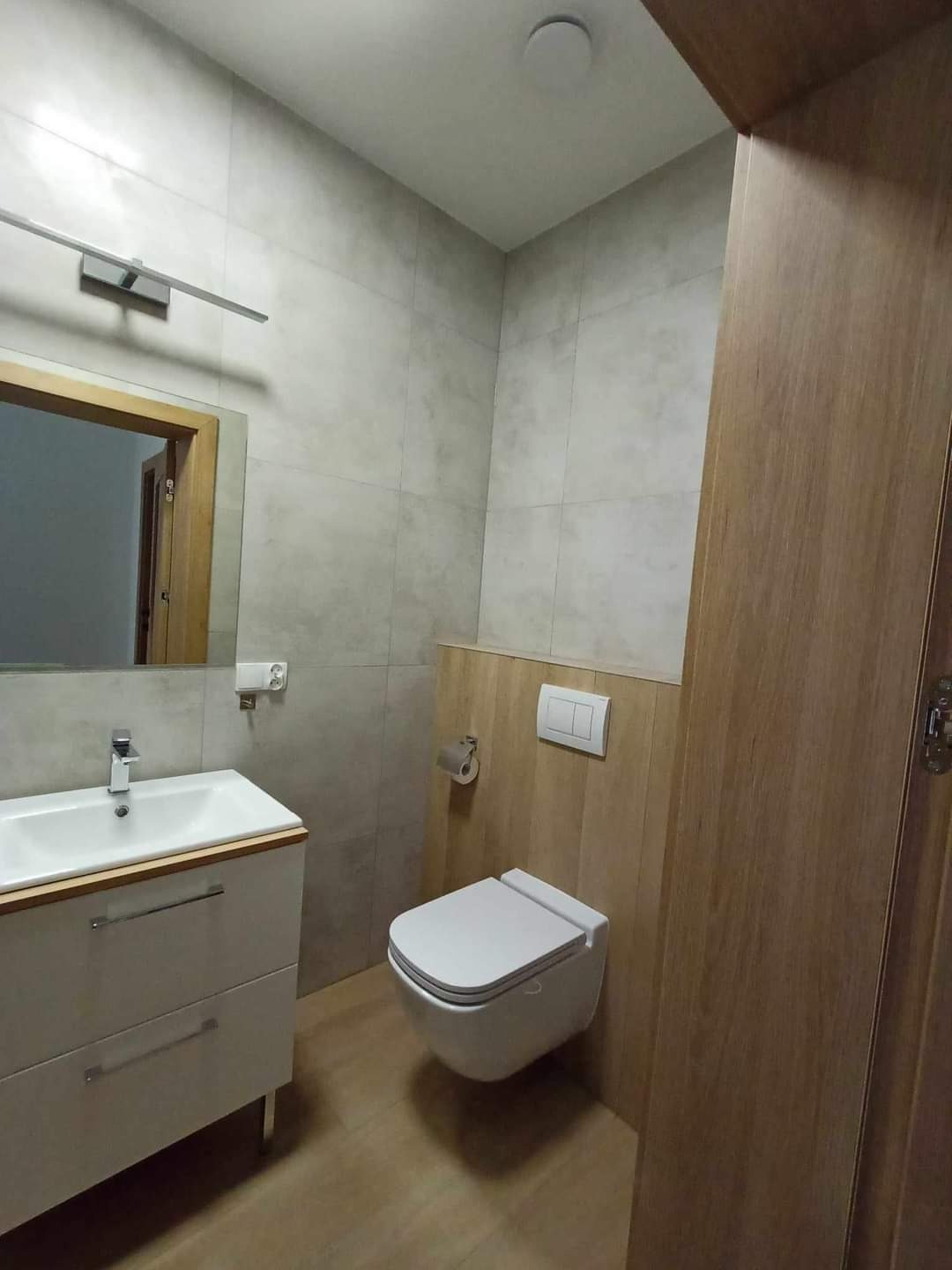 Nowoczesna łazienka z podwieszaną toaletą, szarą glazurą i drewnianymi akcentami, widok na umywalkę z szafką i lustro.