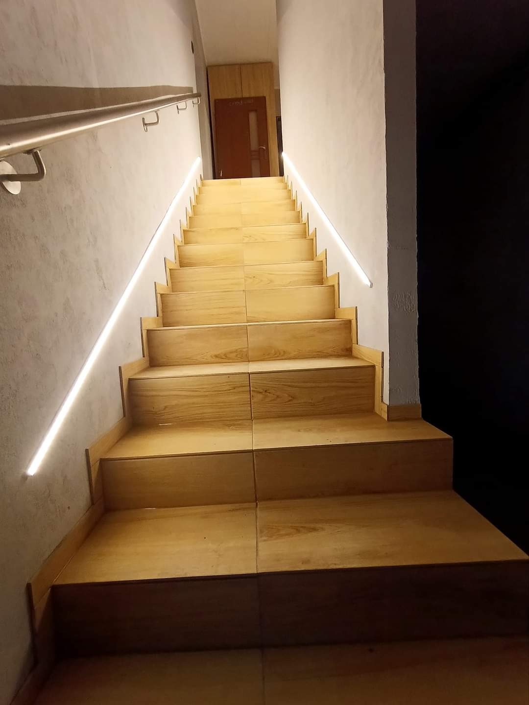 Schody wyłożone płytkami imitującymi drewno, z podświetleniem LED wzdłuż ściany i metalową poręczą, prowadzące do drzwi.
