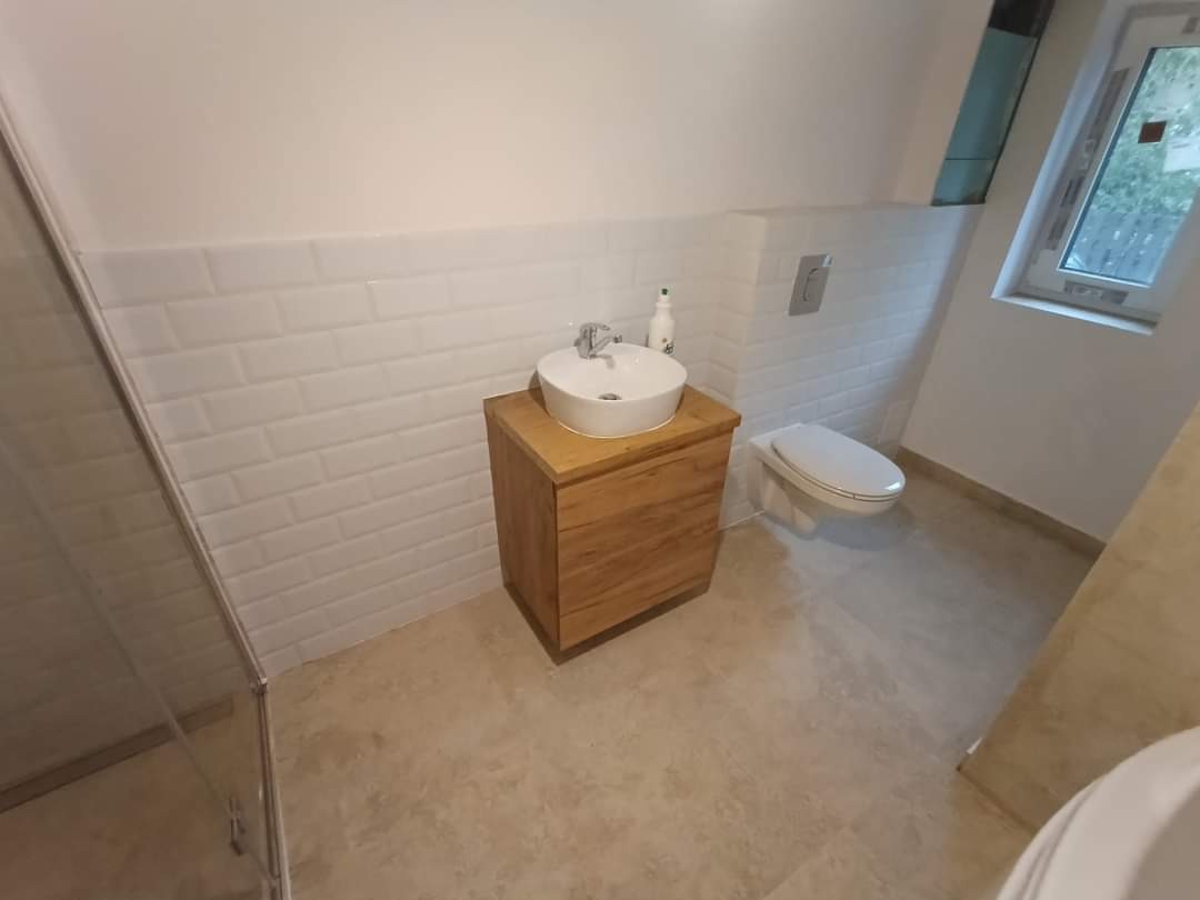 Jasna łazienka po remoncie z drewnianą szafką pod umywalką, białą umywalką nablatową, toaletą podwieszaną i ścianami wyłożonymi białymi, prostokątnymi płytkami imitującymi cegłę.