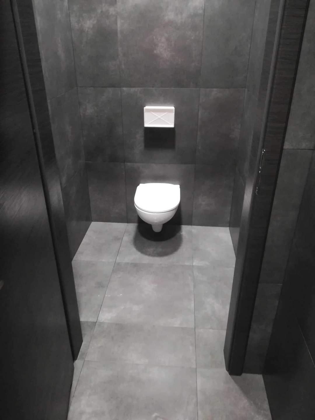 Wnętrze łazienki z szarymi płytkami na ścianach i podłodze, widok na toaletę i podajnik na papier toaletowy z perspektywy wejścia przez otwarte drzwi.