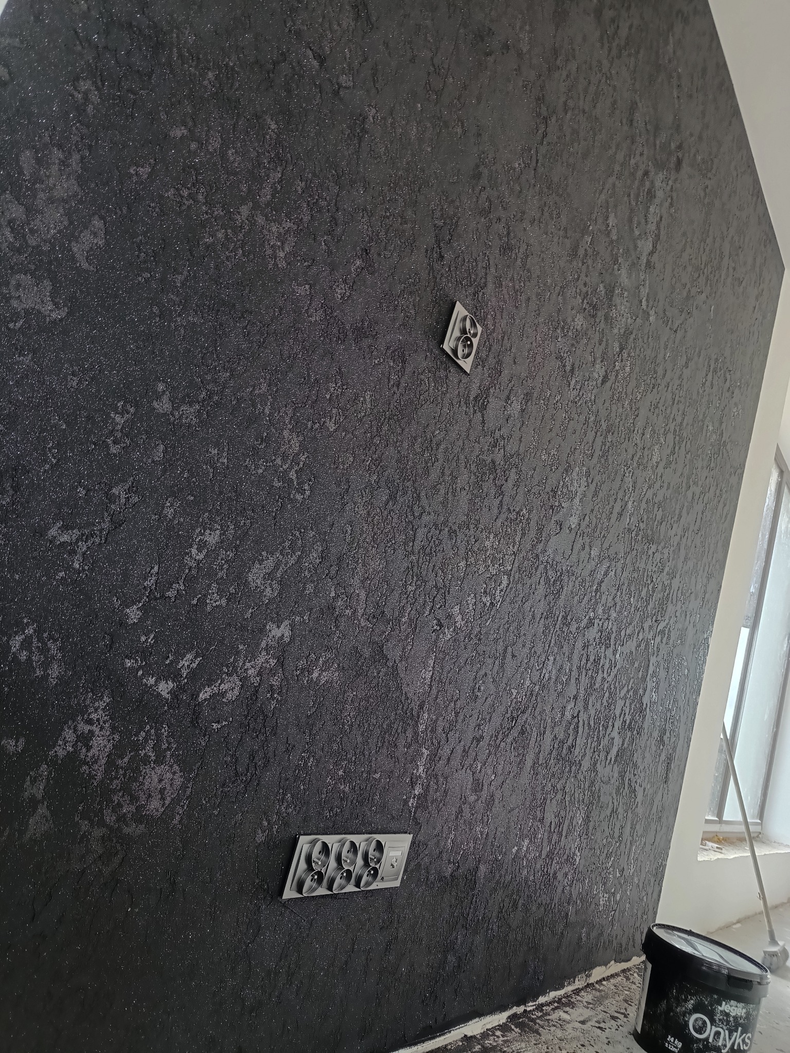 Czarna ściana z dekoracyjną fakturą, widoczne gniazdka elektryczne i puszka farby 'Onyks' przy podłodze.