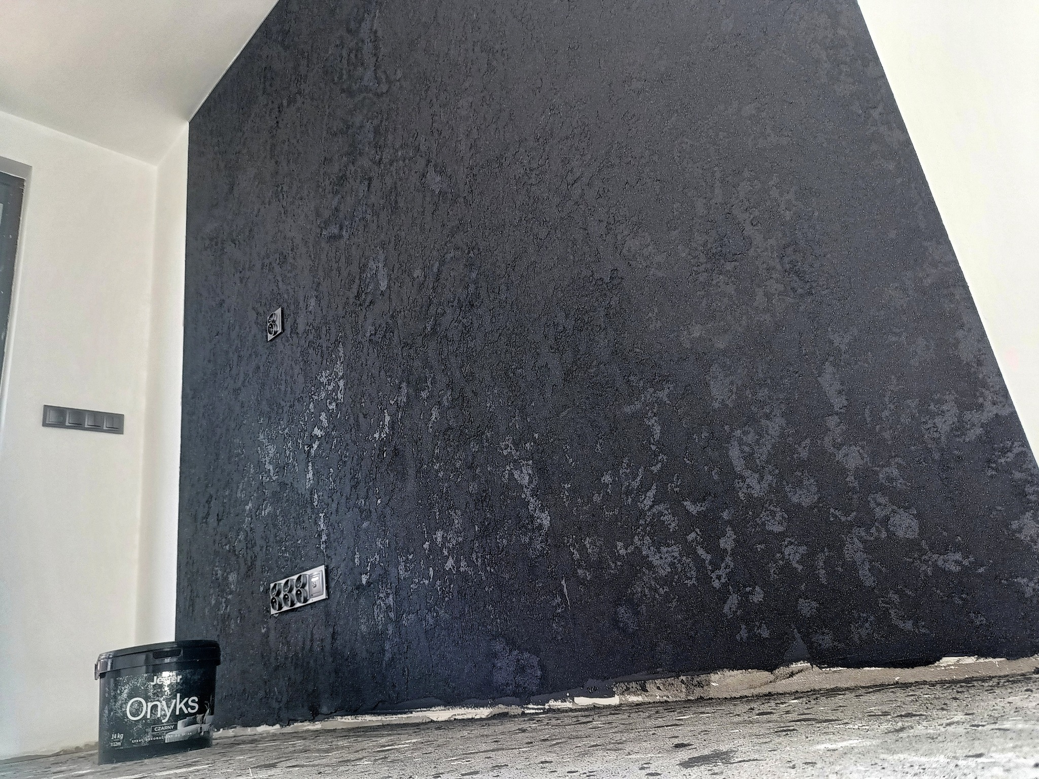 Ściana z fakturą w odcieniach czerni i szarości, z widocznymi gniazdkami elektrycznymi, na podłodze stoi wiadro z farbą 'Onyks', widoczne ślady farby na podłodze.