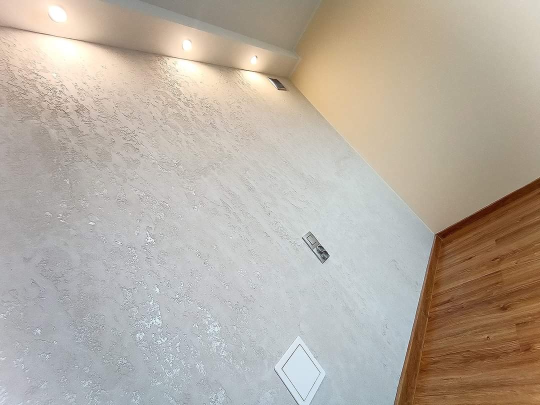 Ściana z dekoracyjną, srebrzystą fakturą w Słupcy, widoczne oświetlenie sufitowe punktowe, drewnopodobna podłoga oraz biały panel dostępowy.