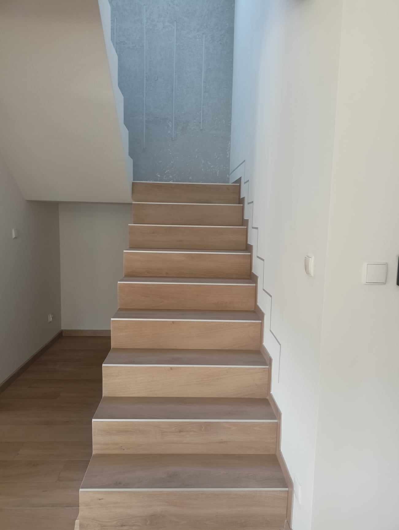Schody wykończone panelami drewnopodobnymi z białymi listwami, na tle białej ściany i szarej ściany o surowym wykończeniu.