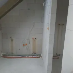 Surowy stan instalacji wodnej i grzewczej w pomieszczeniu z bloków betonowych, widoczne rury rozprowadzone po podłodze i wyprowadzenia ze ściany.