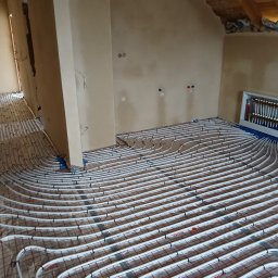 ART-INSTALBUD Artur Popowicz - Rozłożone rury ogrzewania podłogowego na izolacji z siatką, widoczny rozdzielacz w białej obudowie, surowy stan ścian i poddasza.